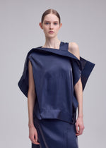 A woman with fair skin and straight brown hair tied back wears the ISSEY MIYAKE MERCURY collection: a shiny, asymmetrical navy blue metallic one-shoulder top with a matching skirt, standing against a plain light grey background.
