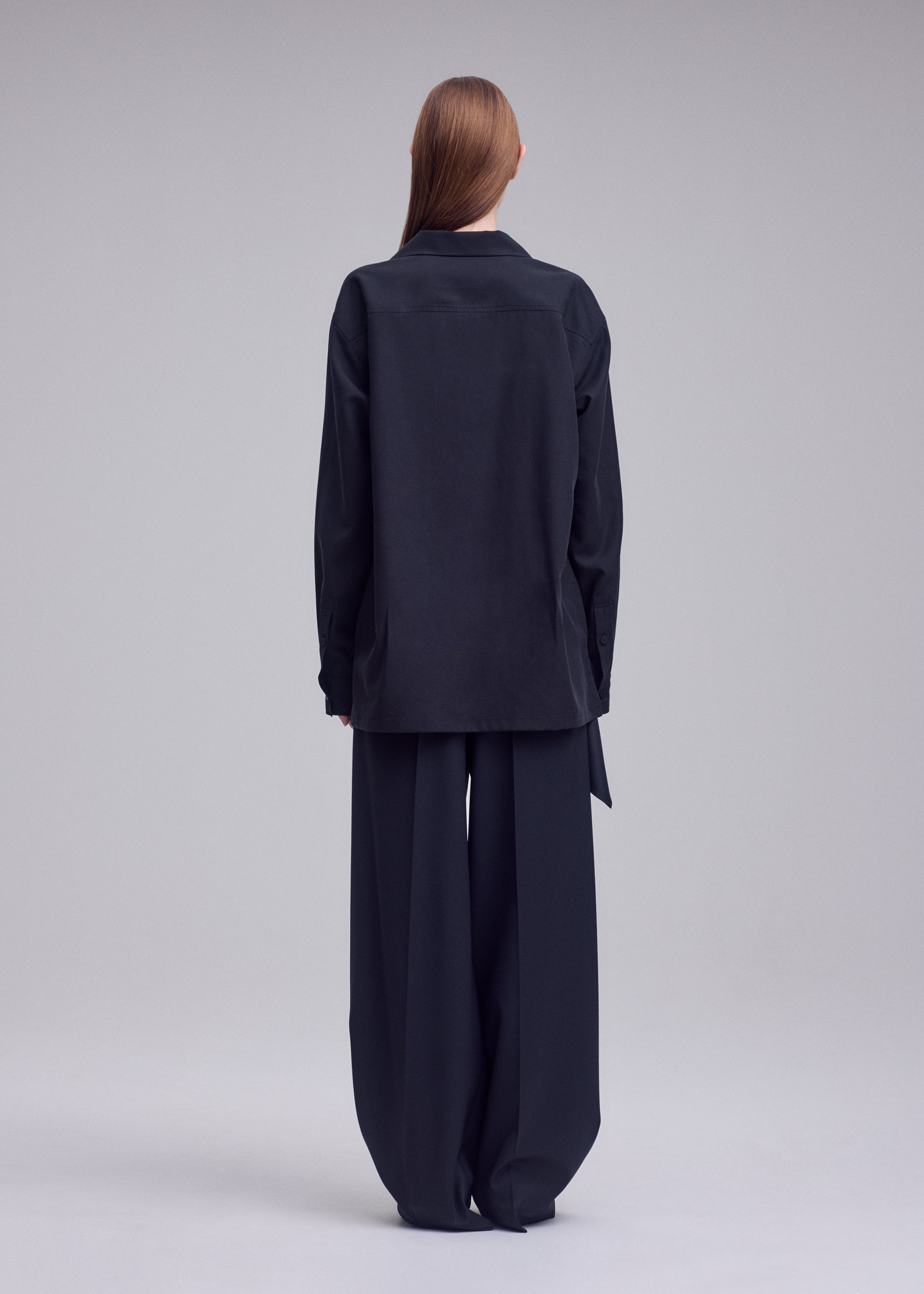 Zoomed view A person with long hair stands facing away, wearing the HANDS OF LOVER loose black open-collar shirt crafted from poplin fabric and wide-leg black pants by ISSEY MIYAKE against a plain gray background.