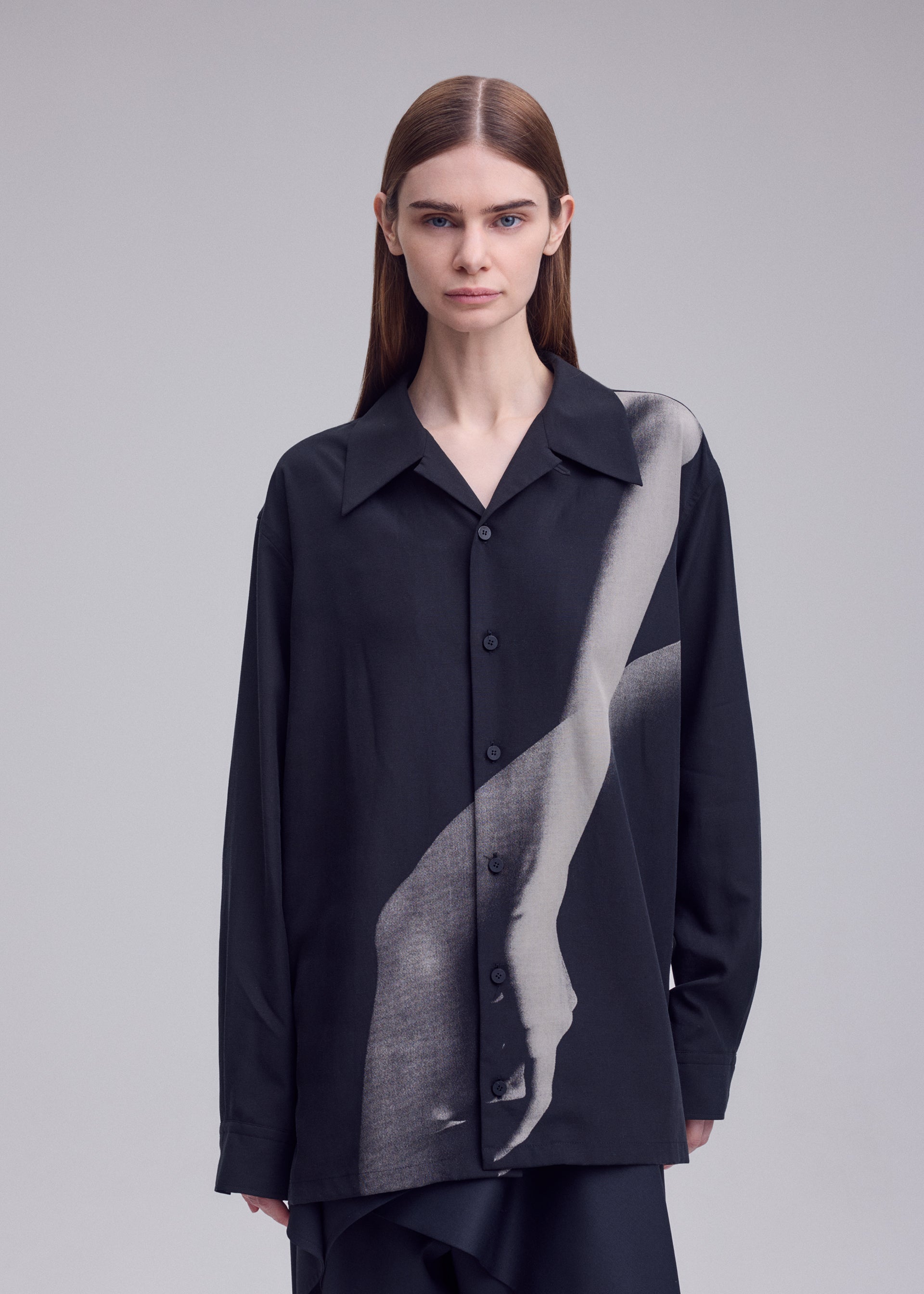 Zoomed view A person with straight brown hair wears the ISSEY MIYAKE HANDS OF LOVER shirt—an open-collar black poplin piece adorned with a grayscale print of an arm and hand across the front—standing against a light gray background.