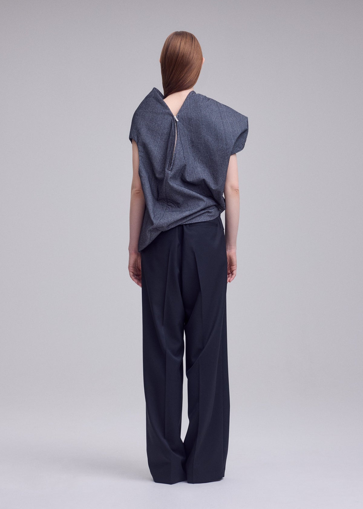 A person stands facing away, wearing the REFLECTION loose gray reflective top with a visible back zipper from ISSEY MIYAKE, paired with wide-leg black trousers. Their straight brown hair falls down their back, and the background is plain and light-colored. view 5 of 7
