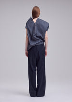 A person stands facing away, wearing the REFLECTION loose gray reflective top with a visible back zipper from ISSEY MIYAKE, paired with wide-leg black trousers. Their straight brown hair falls down their back, and the background is plain and light-colored.