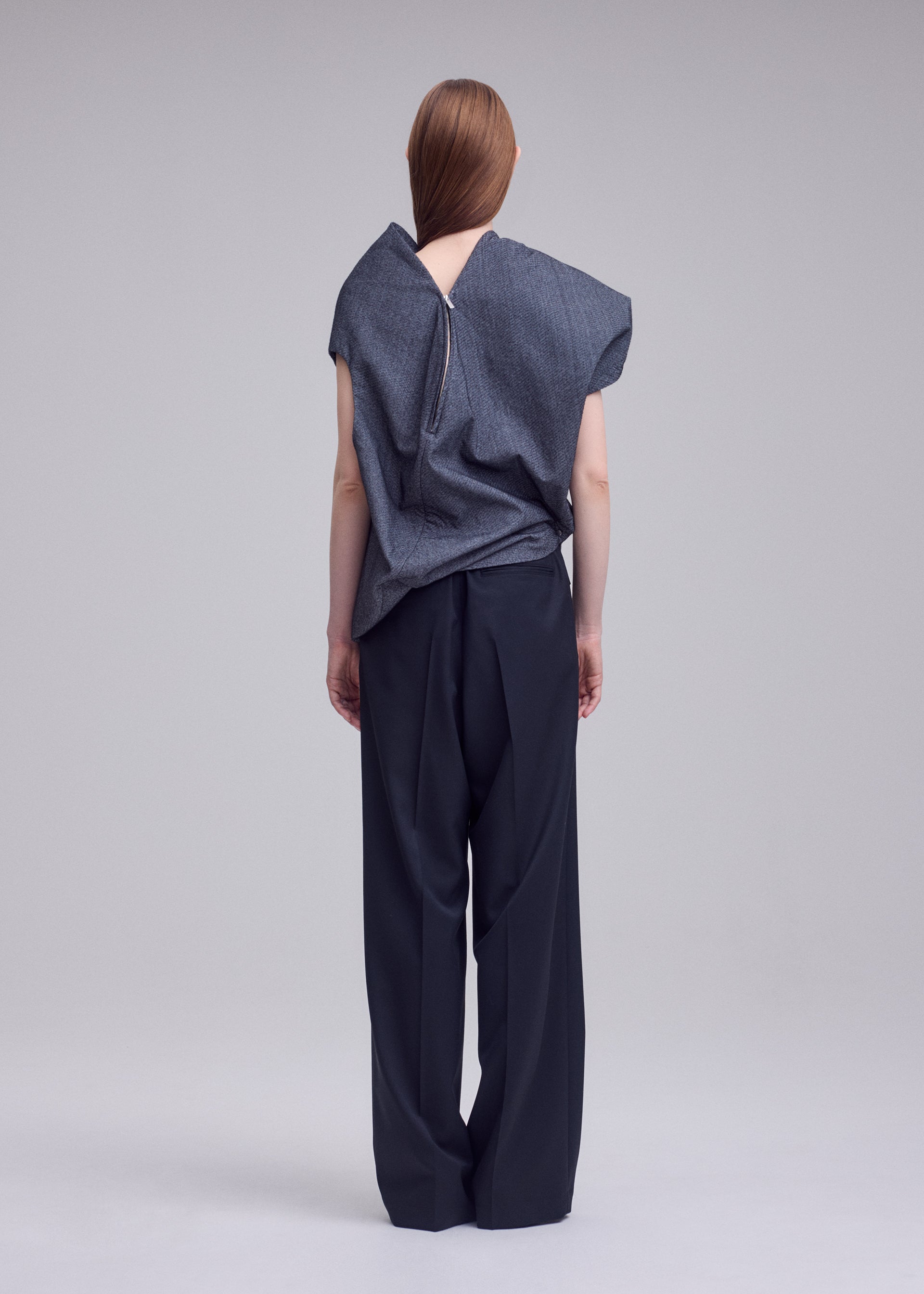 Zoomed view A person stands facing away, wearing the REFLECTION loose gray reflective top with a visible back zipper from ISSEY MIYAKE, paired with wide-leg black trousers. Their straight brown hair falls down their back, and the background is plain and light-colored.