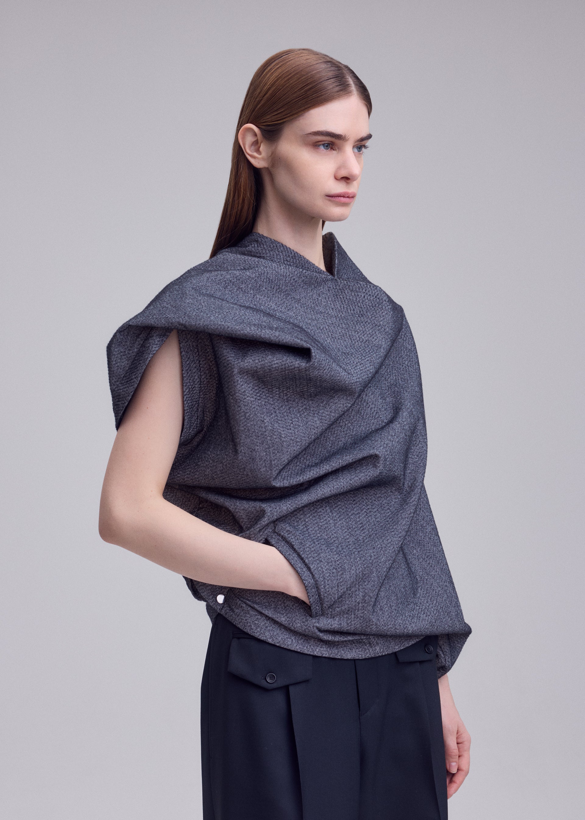 Zoomed view A person with straight, brown hair wears the REFLECTION top by ISSEY MIYAKE—a gray, draped, sleeveless piece crafted from glowing fabric—paired with black pants and stands with one hand in a pocket against a plain background.