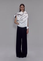 A person stands against a plain gray background wearing ISSEY MIYAKE’s REFLECTION—a shiny, draped silver top—paired with loose black trousers. Their expression is neutral, with one hand in their pocket.