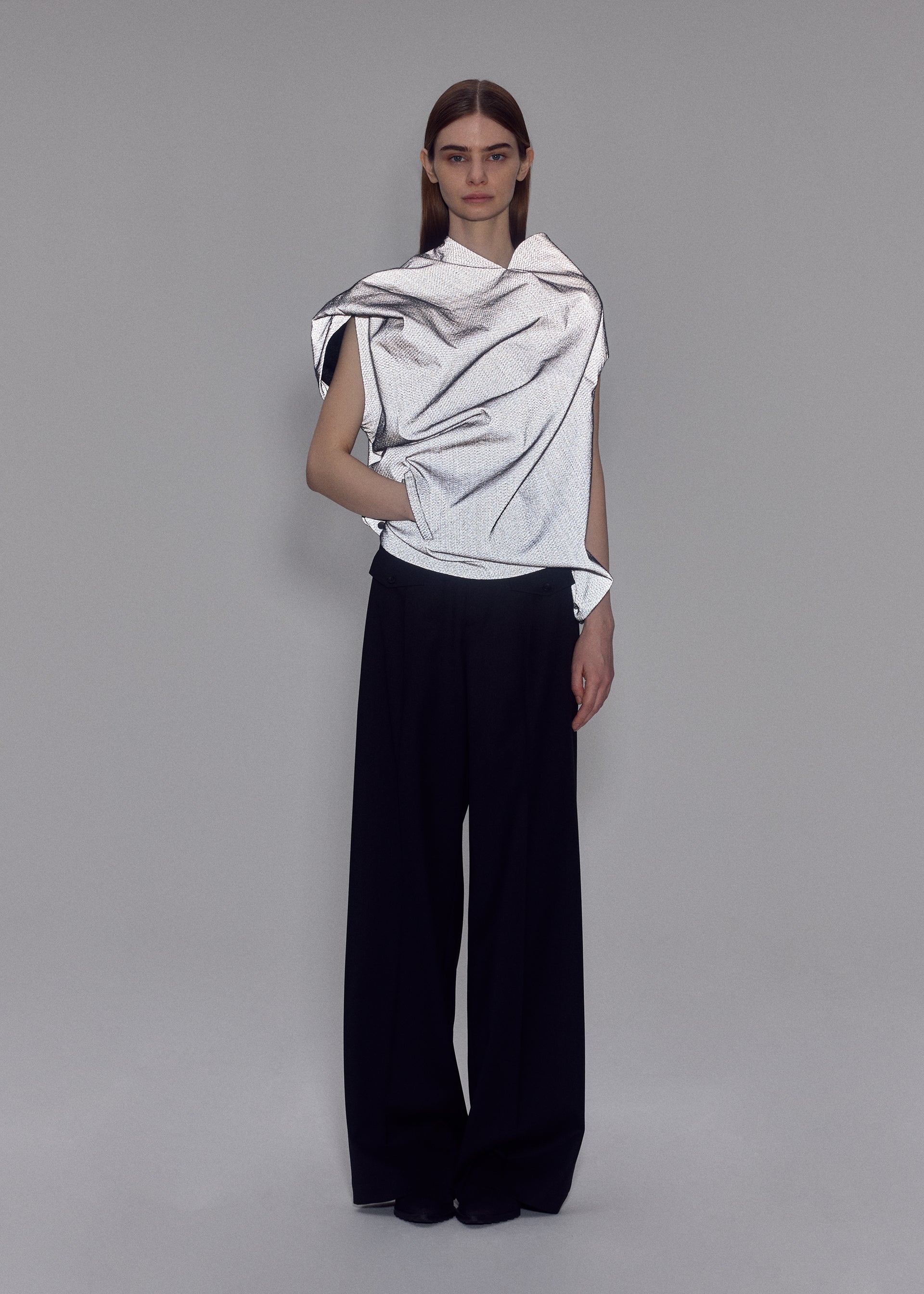 Zoomed view A person stands against a plain gray background wearing ISSEY MIYAKE’s REFLECTION—a shiny, draped silver top—paired with loose black trousers. Their expression is neutral, with one hand in their pocket.