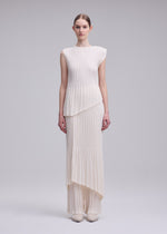 A woman stands against a light gray background wearing ISSEY MIYAKE's UNFURLING PLEATS—a sleeveless ivory top and matching long skirt with diagonal Keshi pleats and layered hems. She has straight brown hair pulled back and wears white shoes.