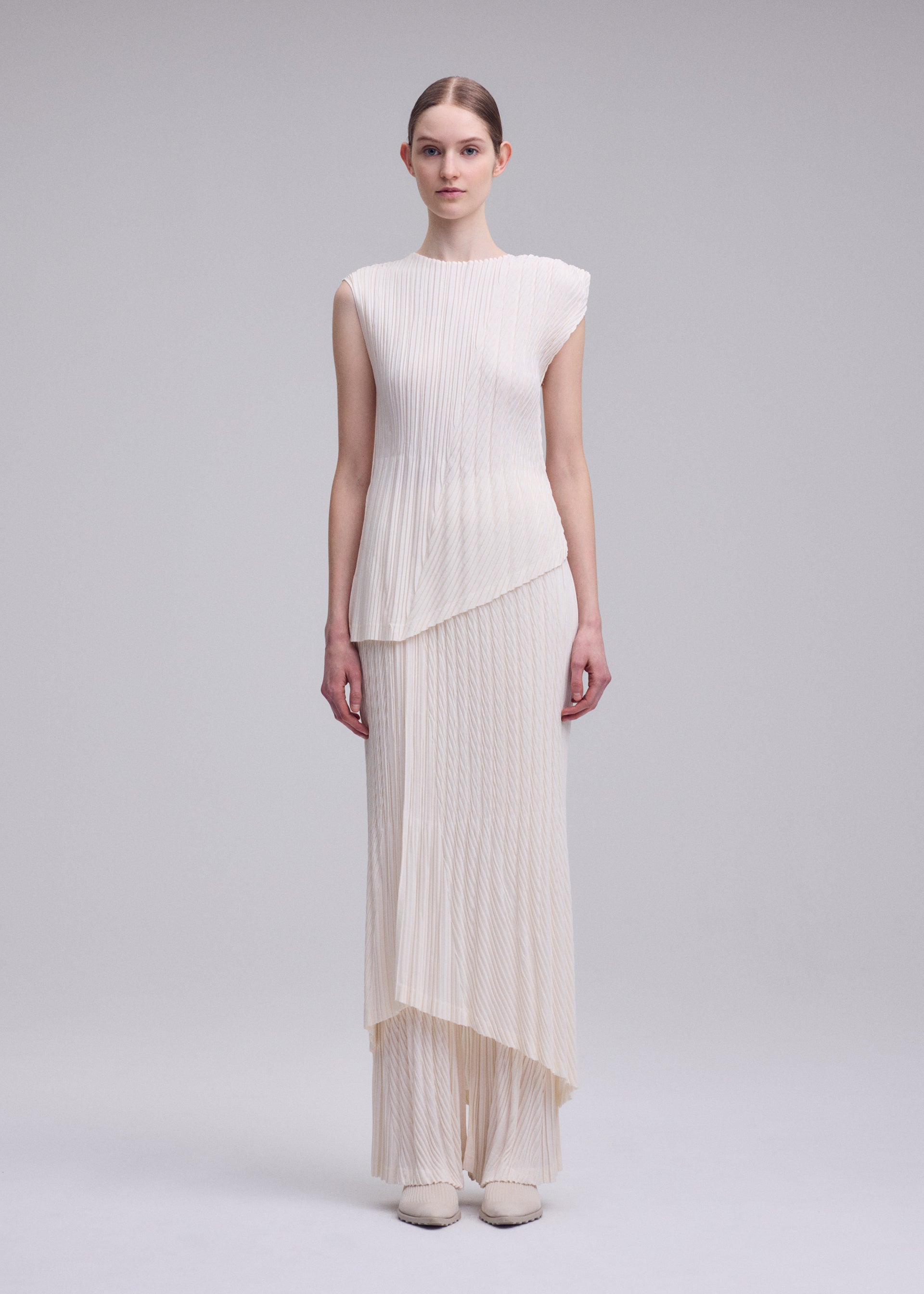 Zoomed view A woman stands against a light gray background wearing ISSEY MIYAKE's UNFURLING PLEATS—a sleeveless ivory top and matching long skirt with diagonal Keshi pleats and layered hems. She has straight brown hair pulled back and wears white shoes.