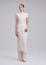 In a minimal studio, a person wears the ISSEY MIYAKE UNFURLING PLEATS dress in ivory, featuring an asymmetrical hem and sleeveless, pleated polyester design, exuding modern elegance as they gaze off to the side.