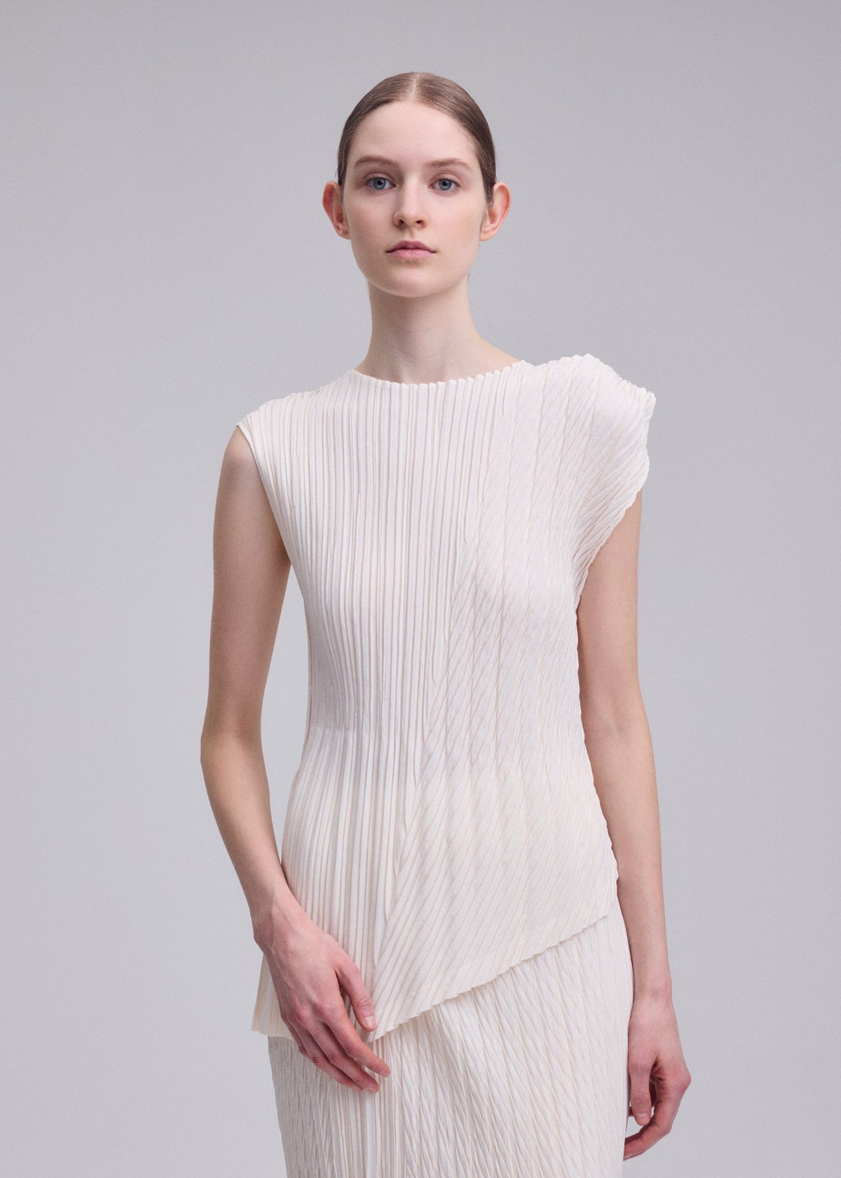 A woman with light skin and brown hair tied back wears ISSEY MIYAKE's UNFURLING PLEATS, a sleeveless white pleated top with an asymmetrical hem, standing against a plain gray background and looking at the camera with a neutral expression. view 7 of 7