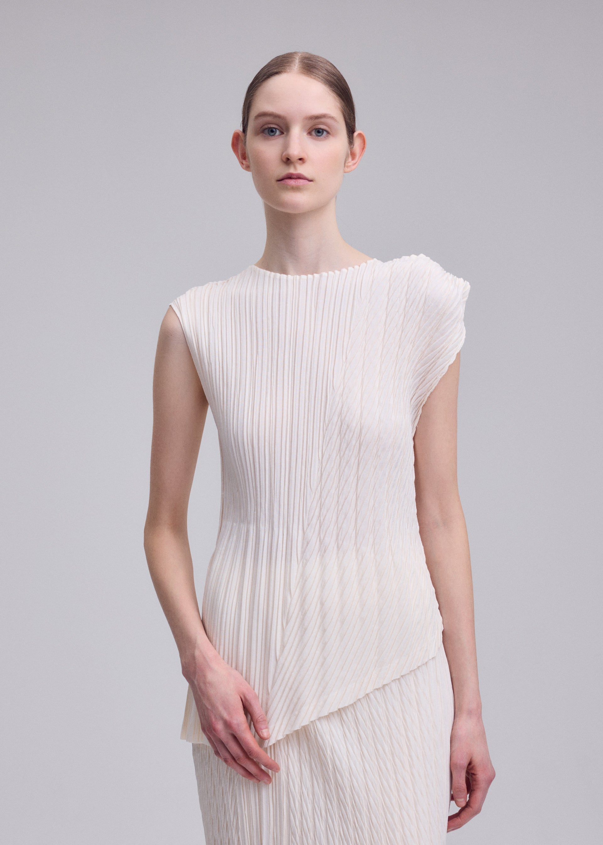 Zoomed view A woman with light skin and brown hair tied back wears ISSEY MIYAKE's UNFURLING PLEATS, a sleeveless white pleated top with an asymmetrical hem, standing against a plain gray background and looking at the camera with a neutral expression.