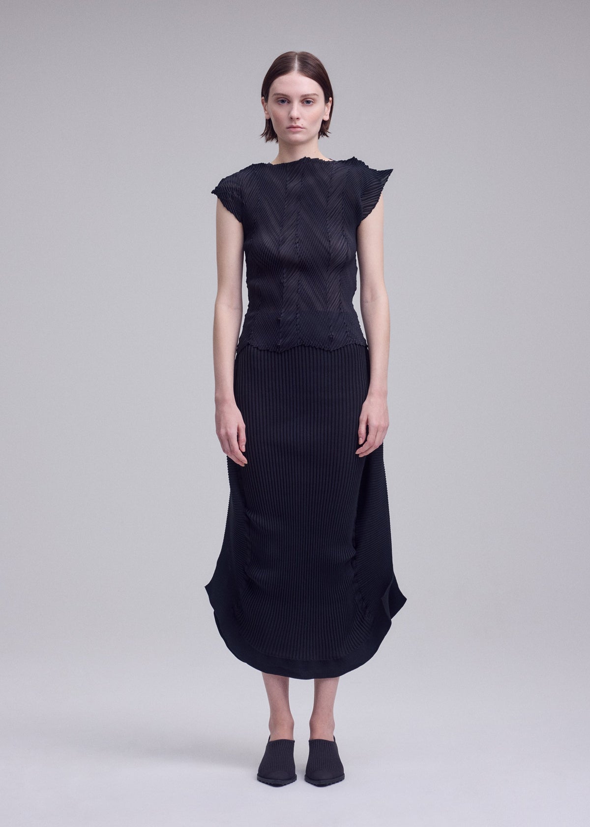A woman stands against a plain gray background wearing the ISSEY MIYAKE ZIGZAG PLEATS black textured French-sleeve top and matching mid-length pleated skirt, paired with black pointed mules. Her arms rest straight at her sides. view 5 of 7