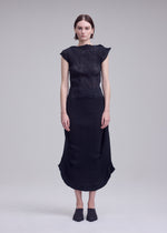 A woman stands against a plain gray background wearing the ISSEY MIYAKE ZIGZAG PLEATS black textured French-sleeve top and matching mid-length pleated skirt, paired with black pointed mules. Her arms rest straight at her sides.