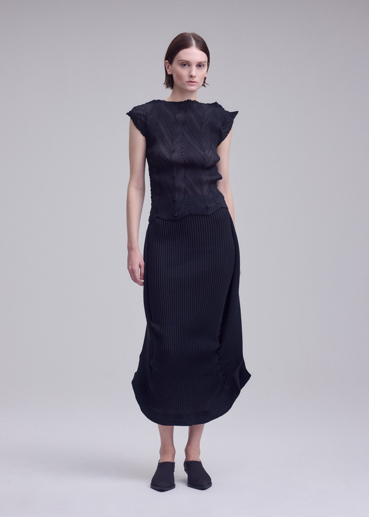 Against a plain background, a person with straight hair wears the ISSEY MIYAKE ZIGZAG PLEATS ensemble—a black, French-sleeve top with textured lines paired with a matching black, calf-length skirt featuring the brand’s signature subtle zigzag pleated pattern. They complete the look with black slip-on shoes. view 6 of 7