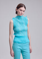 A person with straight brown hair, dressed in the ISSEY MIYAKE ZIGZAG PLEATS high-neck sleeveless top with a zigzag hem and matching turquoise pants, stands against a plain light gray background.