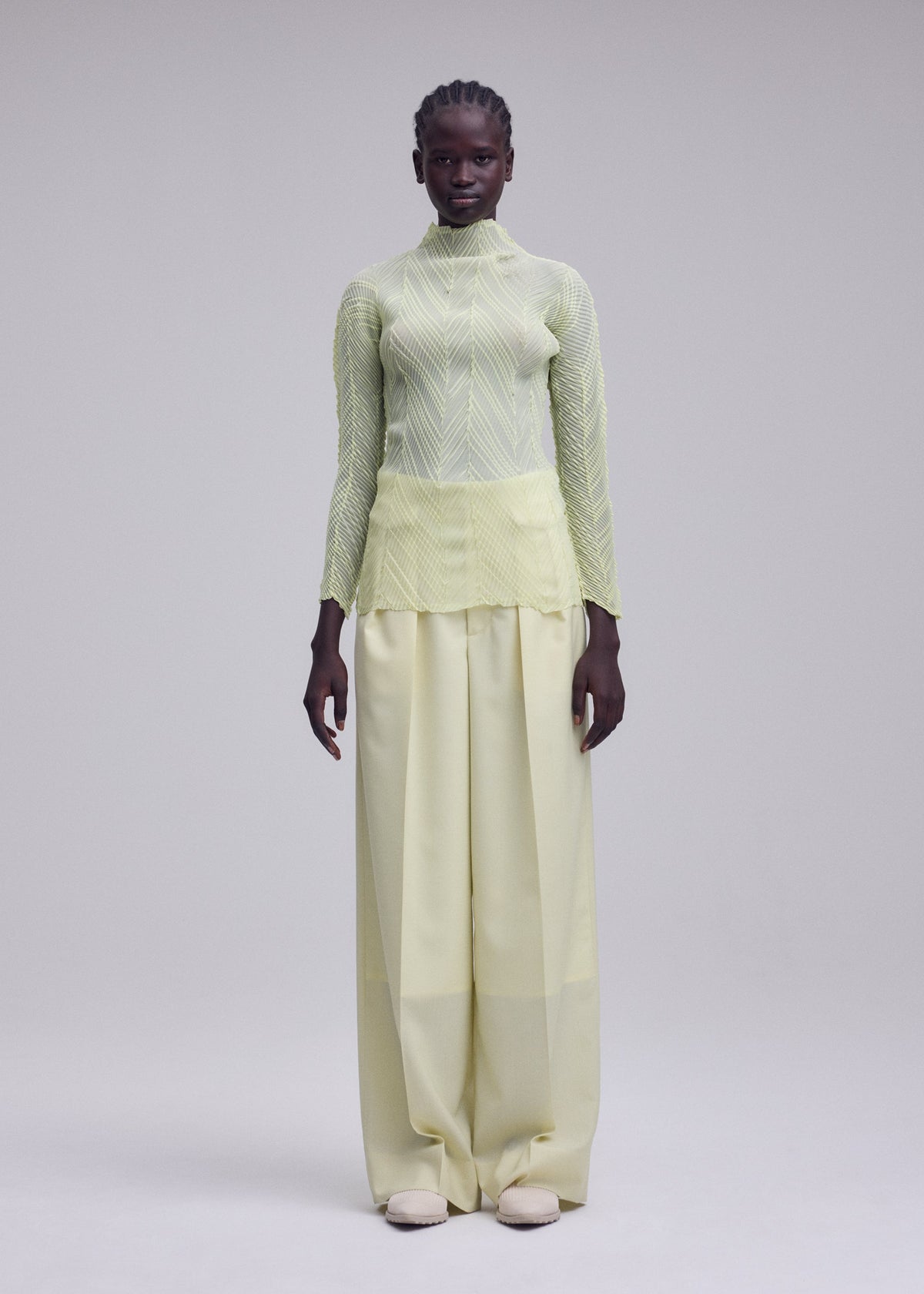 A person stands facing forward against a plain light gray background, wearing the ISSEY MIYAKE ZIGZAG PLEATS: a sheer polyester, light yellow long-sleeve pleated top with a subtle zigzag hem, paired with wide-legged, high-waisted pale yellow pants and white shoes. view 5 of 8