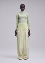 A person stands facing forward against a plain light gray background, wearing the ISSEY MIYAKE ZIGZAG PLEATS: a sheer polyester, light yellow long-sleeve pleated top with a subtle zigzag hem, paired with wide-legged, high-waisted pale yellow pants and white shoes.