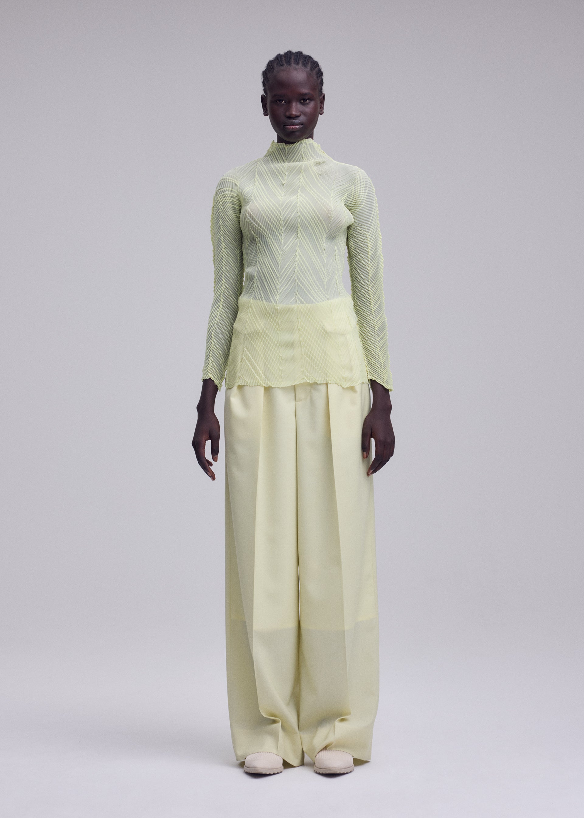 Zoomed view A person stands facing forward against a plain light gray background, wearing the ISSEY MIYAKE ZIGZAG PLEATS: a sheer polyester, light yellow long-sleeve pleated top with a subtle zigzag hem, paired with wide-legged, high-waisted pale yellow pants and white shoes.