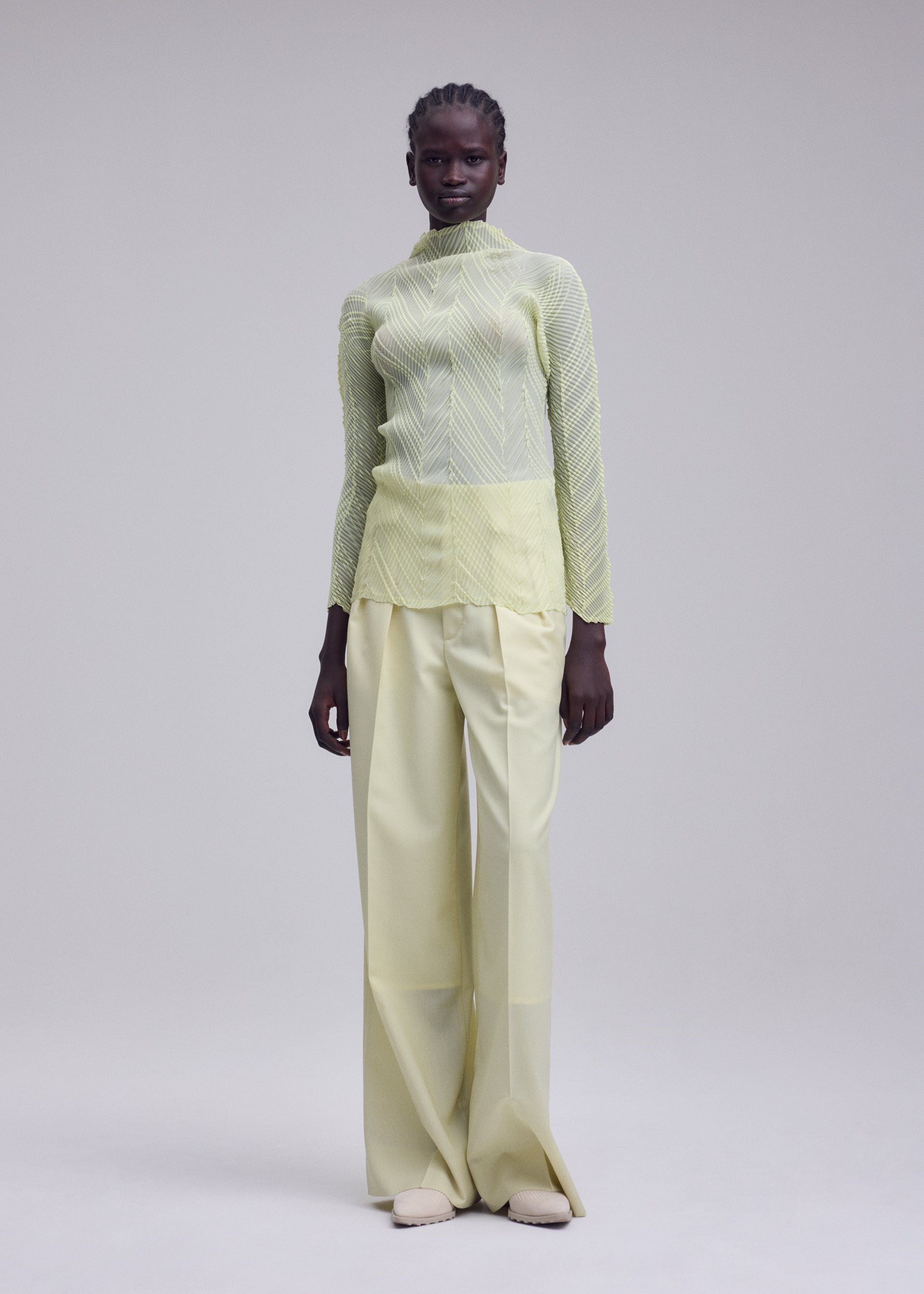 Zoomed view A woman stands against a plain background, wearing the ISSEY MIYAKE ZIGZAG PLEATS sheer polyester light green long-sleeve top with a zigzag hem over a yellow camisole and wide-legged, pale yellow trousers. She has short, braided hair and beige shoes.