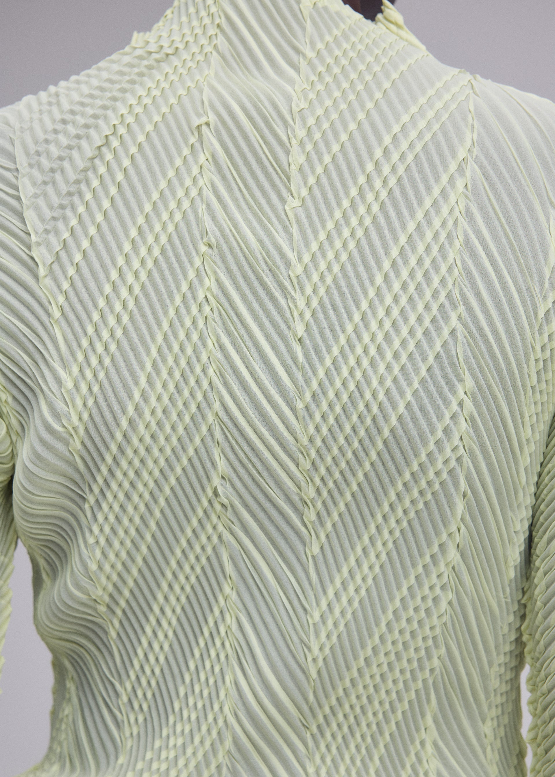 Zoomed view Close-up of the back of the ISSEY MIYAKE ZIGZAG PLEATS top, crafted from light green fabric with structured folds forming a geometric, chevron-inspired pattern and finished with a signature zigzag hem.