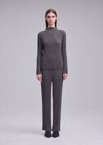 A person stands facing forward in the ISSEY MIYAKE HATCHING PLEATS gray ribbed high-neck long-sleeve top and pants, paired with black pointed shoes against a plain light gray background.