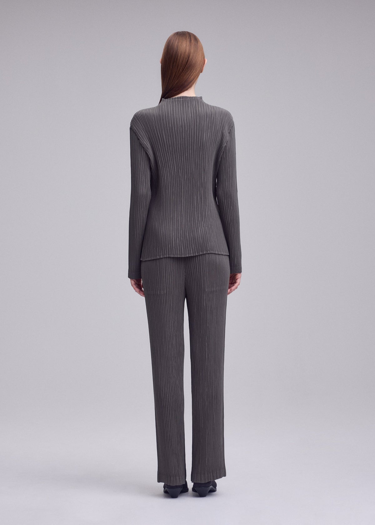 A person with straight brown hair faces away, wearing ISSEY MIYAKE’s HATCHING PLEATS high-neck ribbed gray top and pants with black shoes, set against a plain light gray background. view 5 of 6
