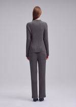 A person with straight brown hair faces away, wearing ISSEY MIYAKE’s HATCHING PLEATS high-neck ribbed gray top and pants with black shoes, set against a plain light gray background.