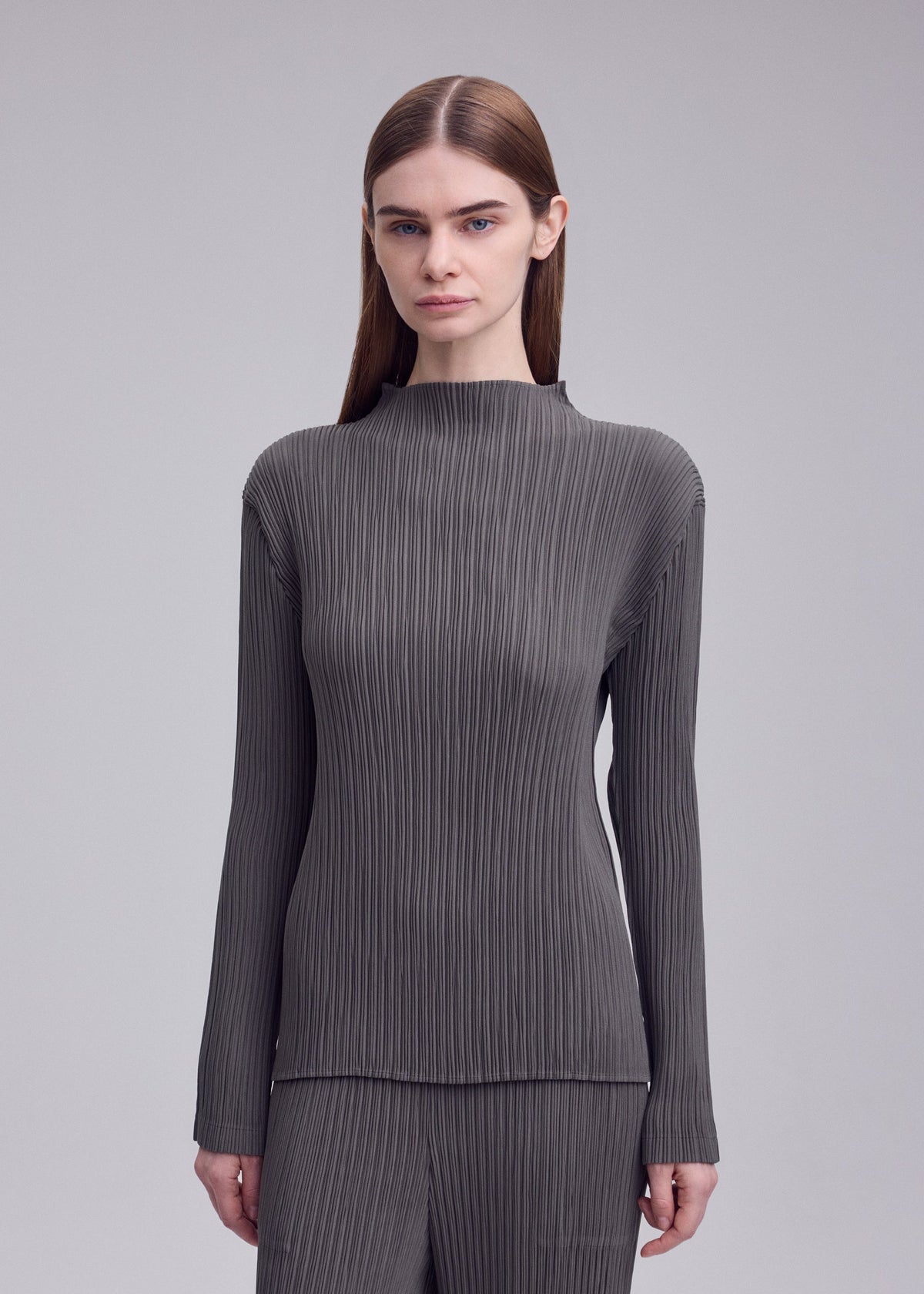 Zoomed view A woman with straight brown hair wears the HATCHING PLEATS set by ISSEY MIYAKE—gray, long-sleeved, high-neck top and matching pants—standing against a plain light gray background.