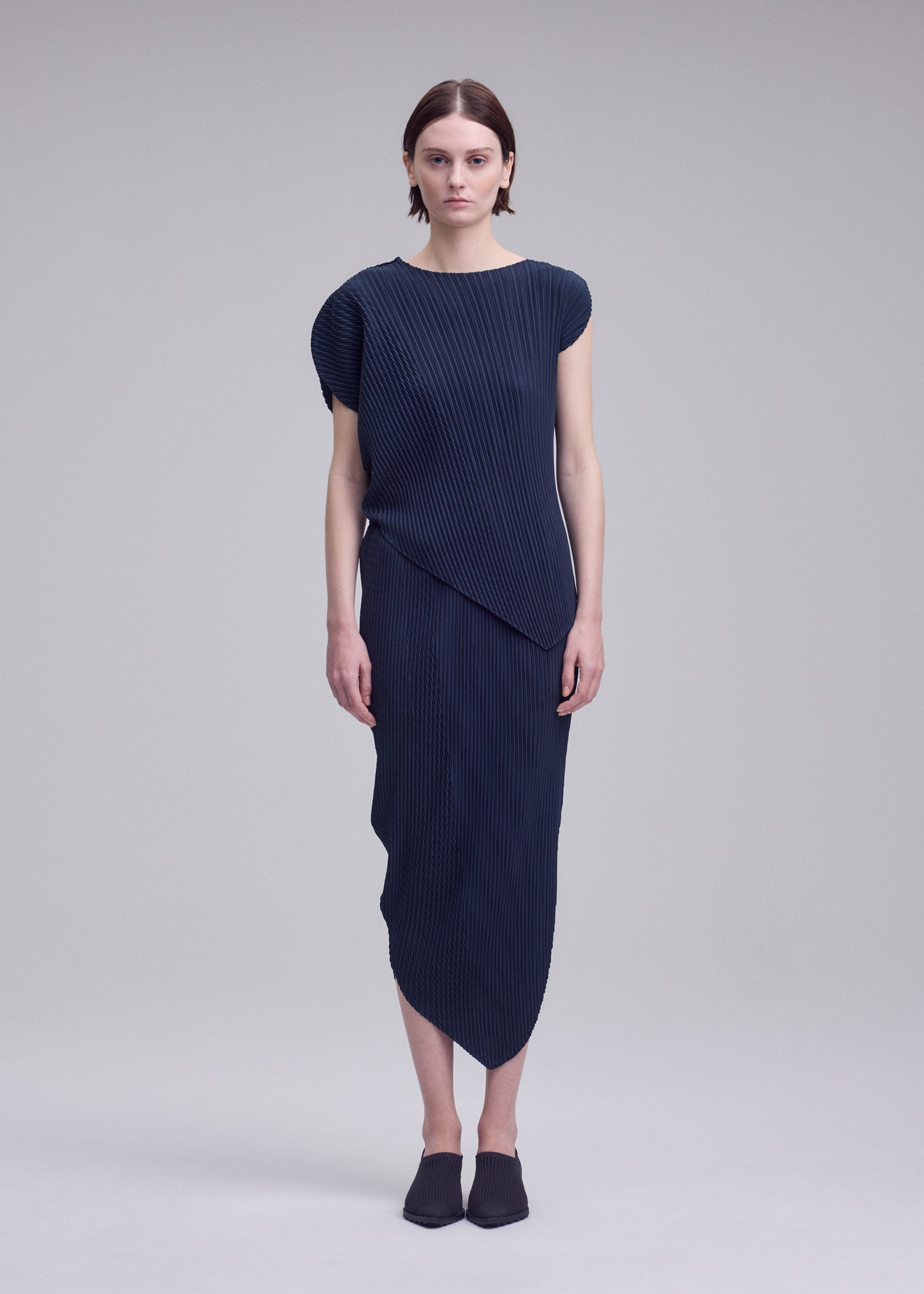 Zoomed view A person stands facing forward against a plain light gray background, wearing the ISSEY MIYAKE LEAF PLEATS navy blue asymmetrical double-pleated short-sleeve dress and black slip-on shoes.