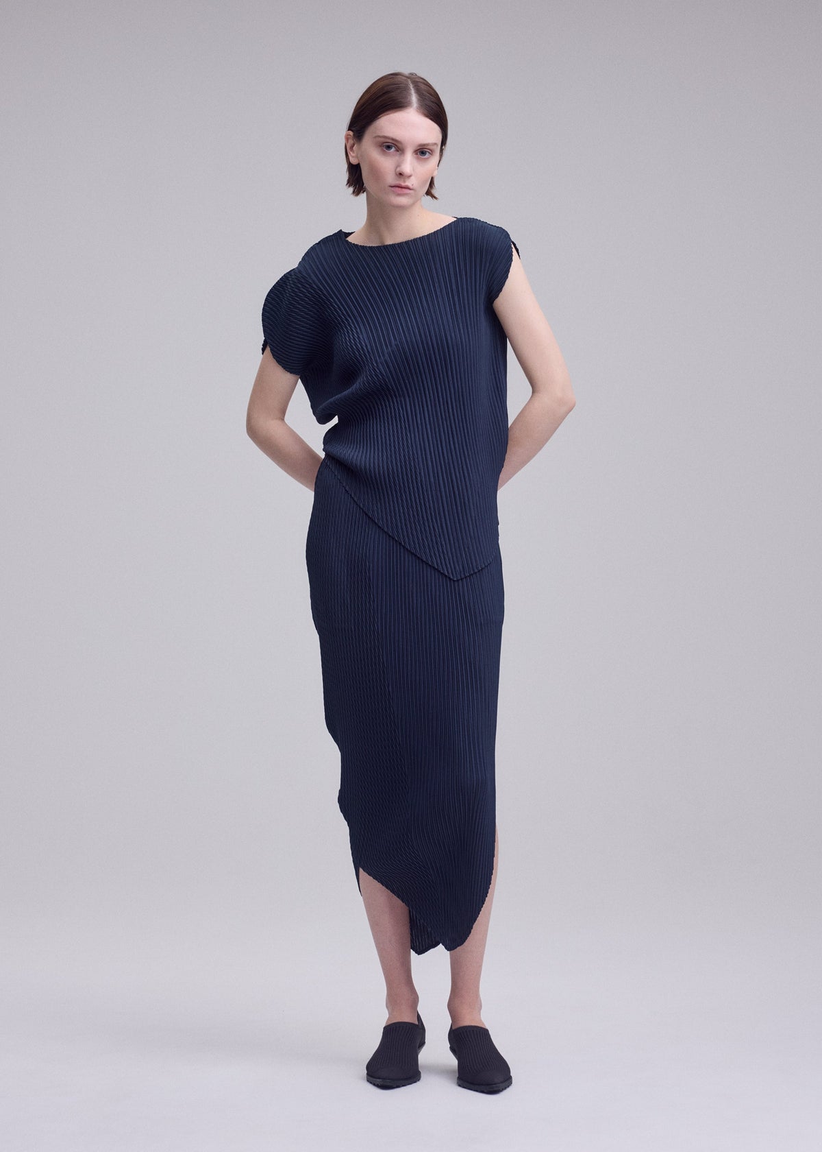 A person with short brown hair models the ISSEY MIYAKE LEAF PLEATS dress—a navy blue, asymmetrical, ribbed piece featuring a subtle leaf silhouette—paired with black shoes and standing against a plain light gray background with their hands behind their back. view 6 of 7