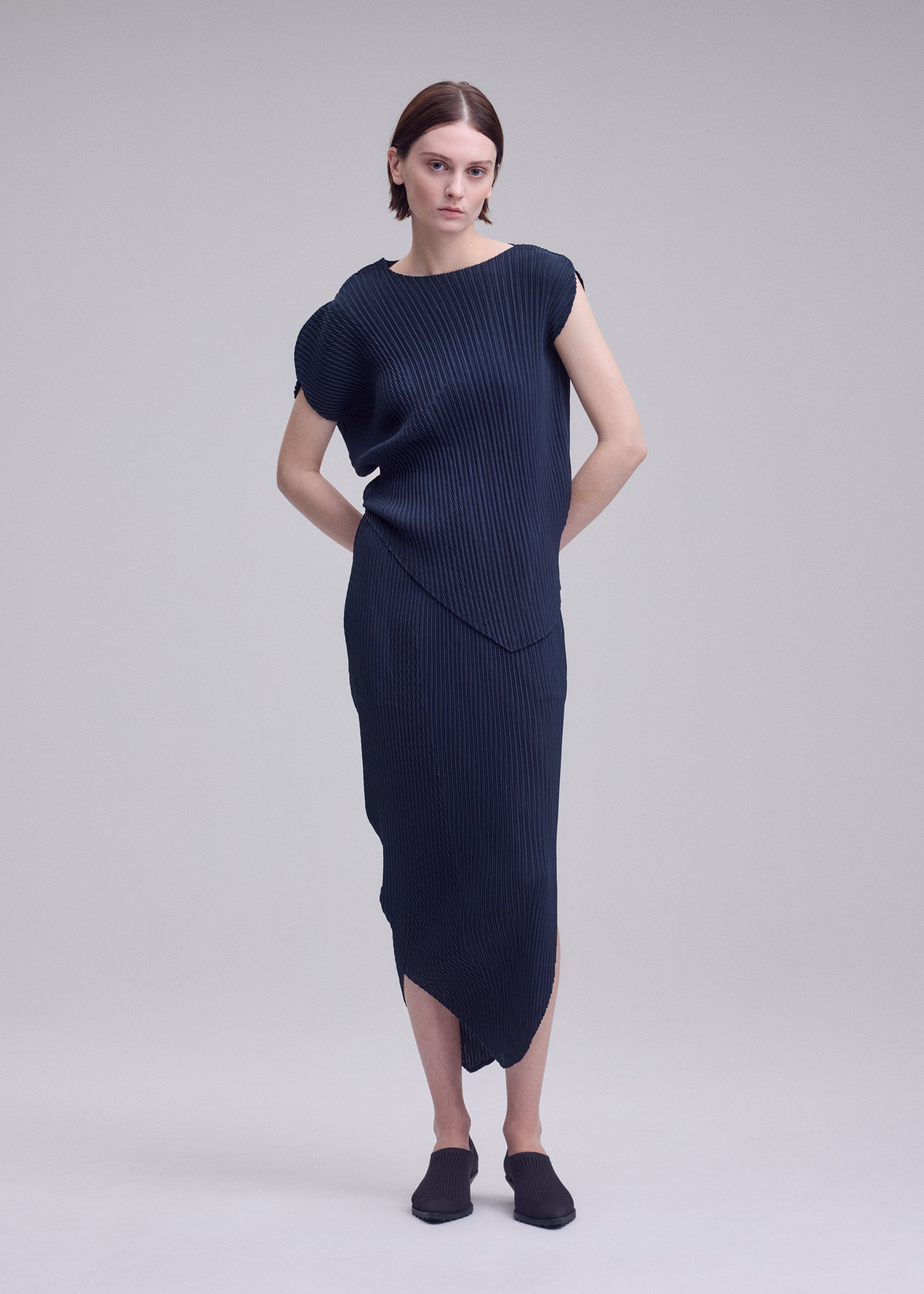 Zoomed view A person with short brown hair models the ISSEY MIYAKE LEAF PLEATS dress—a navy blue, asymmetrical, ribbed piece featuring a subtle leaf silhouette—paired with black shoes and standing against a plain light gray background with their hands behind their back.