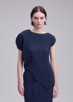 A person with short brown hair wears the LEAF PLEATS dress by ISSEY MIYAKE—a dark blue, asymmetrical, pleated design featuring subtle double-pleating—and stands against a plain light gray background, looking directly at the camera.