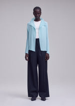 A person stands against a plain light gray background wearing the ISSEY MIYAKE MISTY PLEATS light blue cardigan, crafted from soft fabric, layered over a white top and wide-leg black trousers.