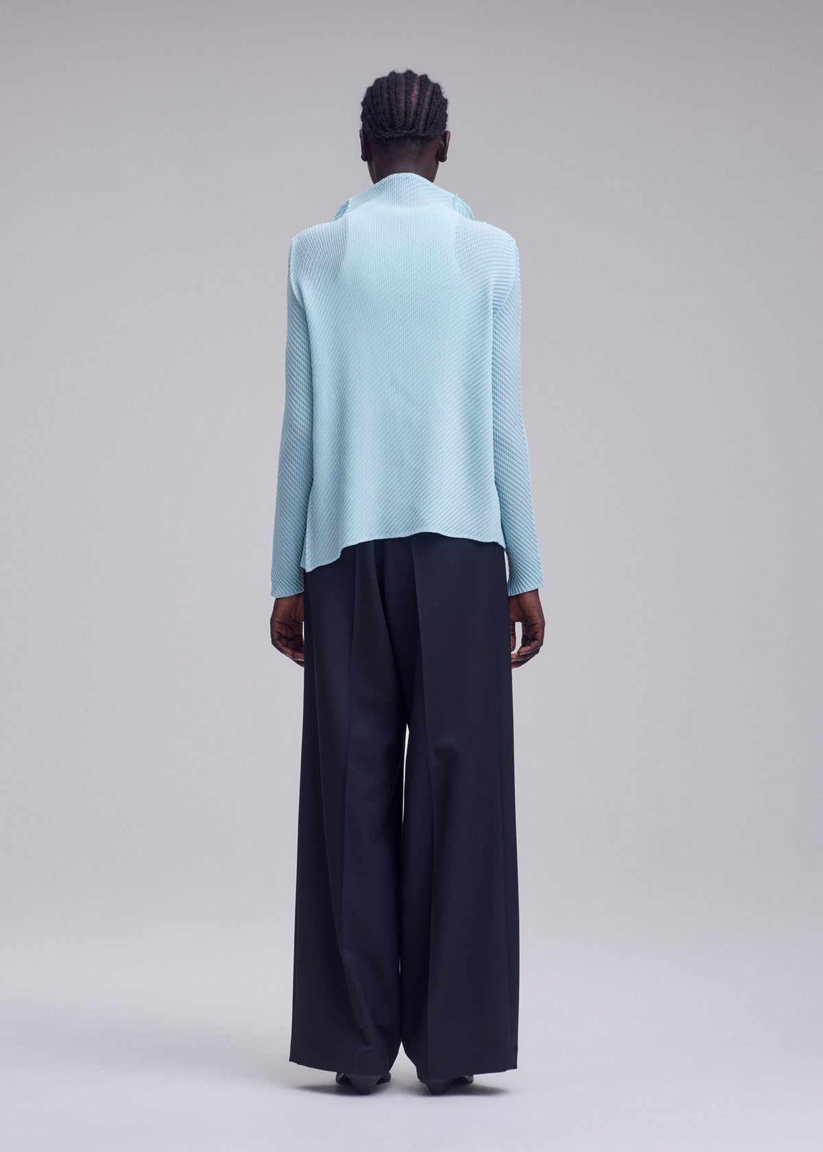 A person stands facing away, wearing the ISSEY MIYAKE MISTY PLEATS light blue long-sleeve top crafted from soft fabric and loose black pants against a plain, light gray background. view 6 of 7
