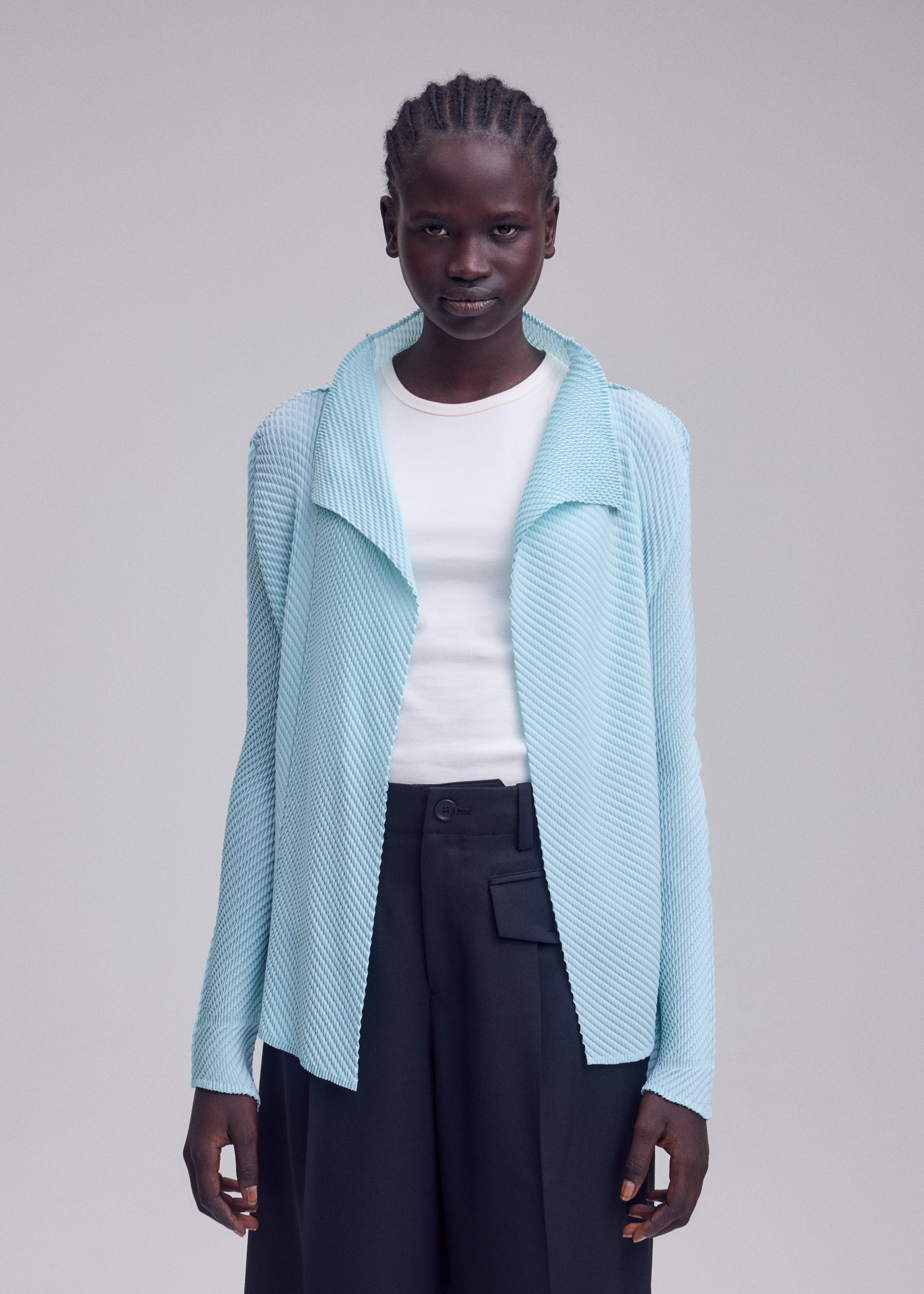 Zoomed view A person wearing the ISSEY MIYAKE MISTY PLEATS light blue pleated cardigan over a white shirt and dark high-waisted trousers stands against a plain background, looking directly at the camera.