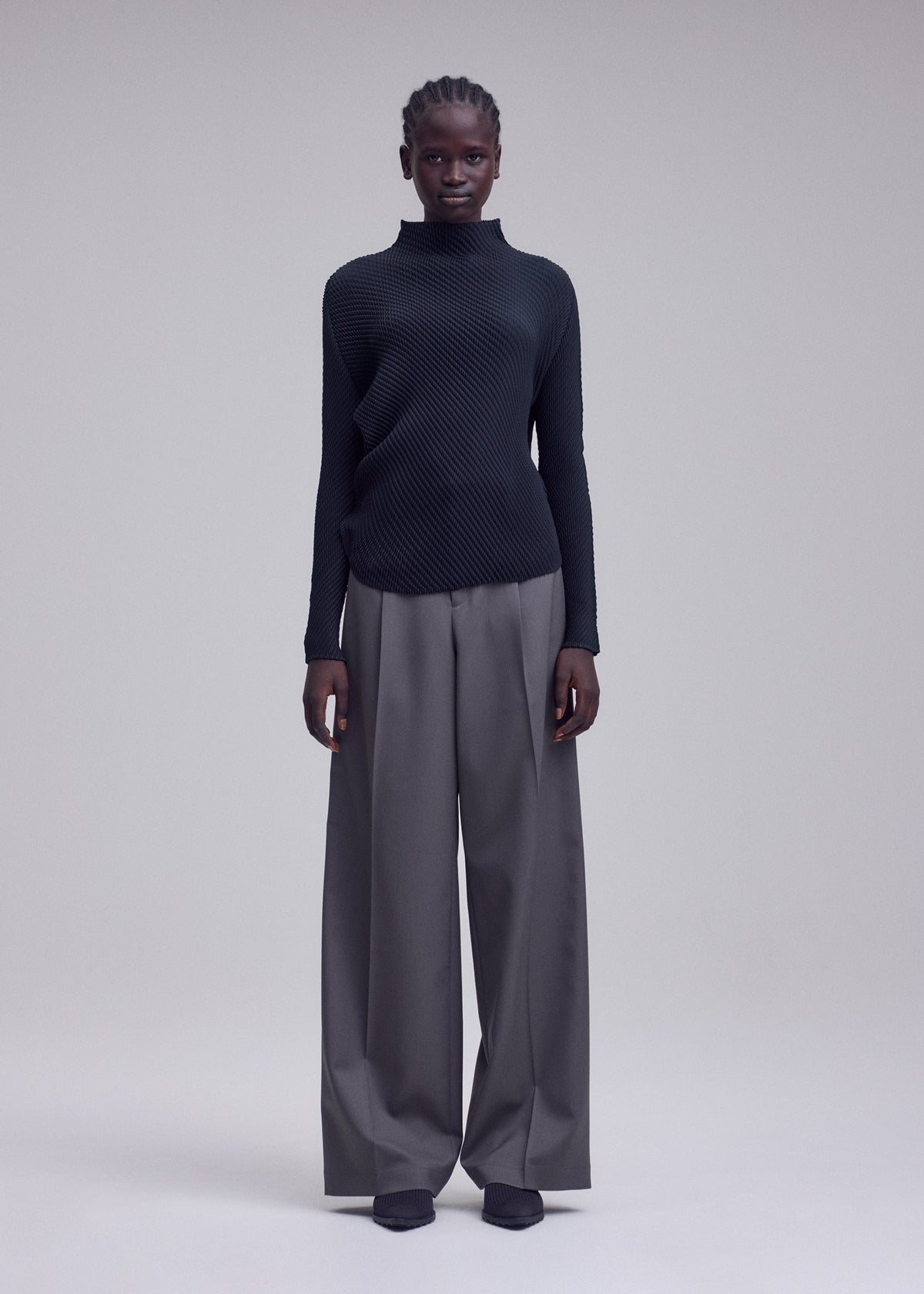 A person stands facing forward, wearing the ISSEY MIYAKE MISTY PLEATS textured black high-neck top and wide-legged gray trousers with dark shoes. The flowy silhouette is complemented by the plain, light-colored background. view 5 of 7