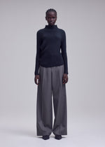 A person stands facing forward, wearing the ISSEY MIYAKE MISTY PLEATS textured black high-neck top and wide-legged gray trousers with dark shoes. The flowy silhouette is complemented by the plain, light-colored background.