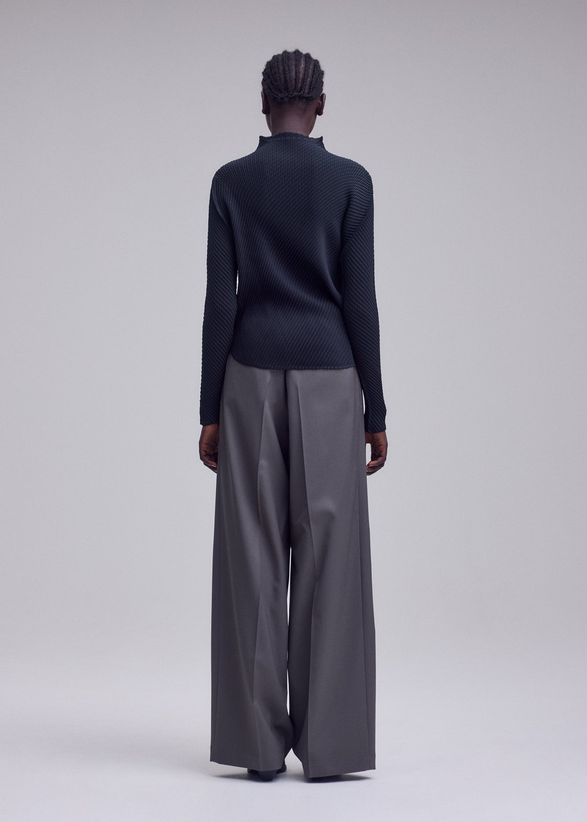 A person stands facing away, wearing the ISSEY MIYAKE MISTY PLEATS top paired with wide-legged gray trousers. The plain, light-colored background accentuates the outfit’s minimalist and modern style, highlighting the signature flowy silhouette. view 6 of 7
