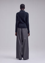 A person stands facing away, wearing the ISSEY MIYAKE MISTY PLEATS top paired with wide-legged gray trousers. The plain, light-colored background accentuates the outfit’s minimalist and modern style, highlighting the signature flowy silhouette.