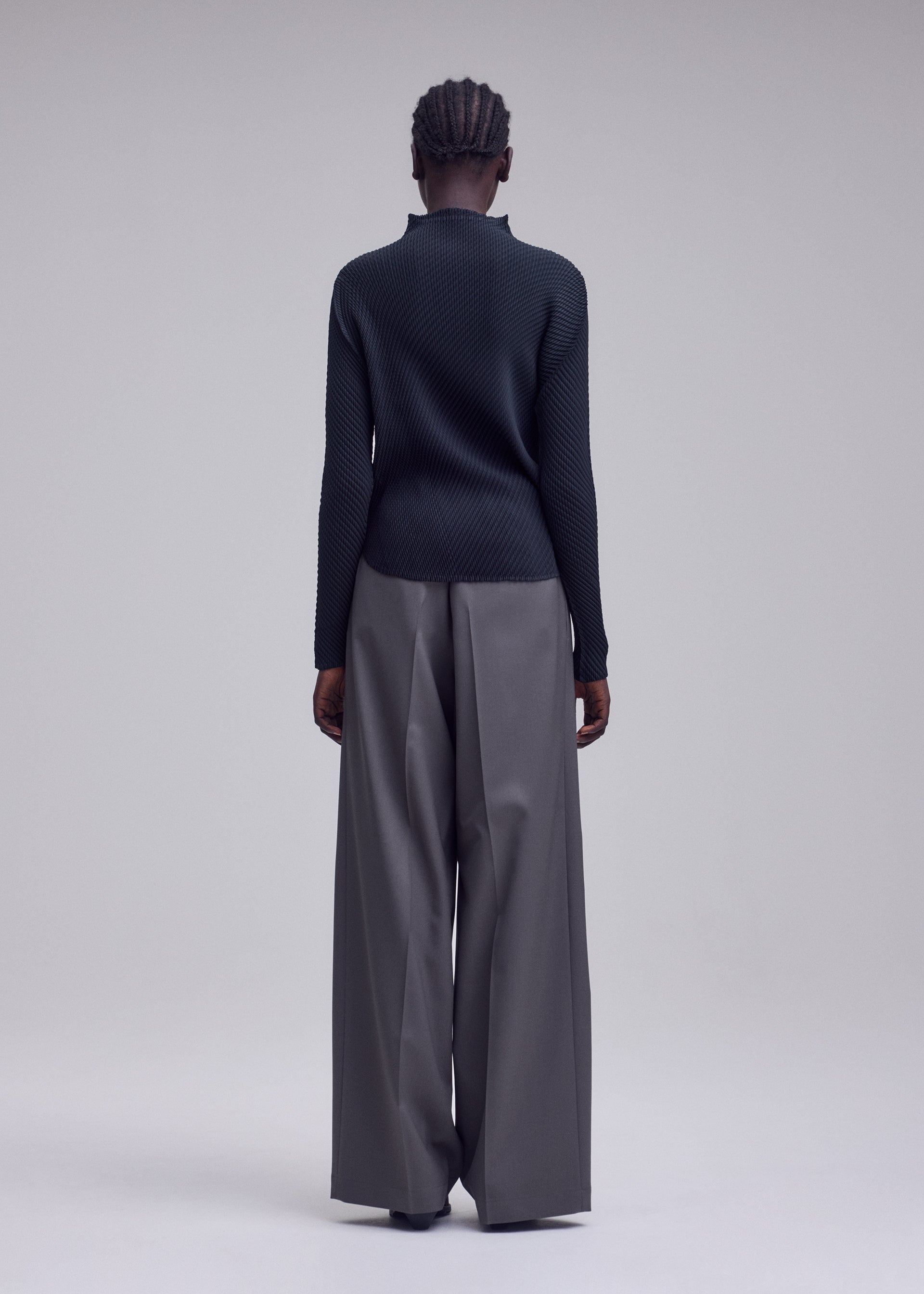 Zoomed view A person stands facing away, wearing the ISSEY MIYAKE MISTY PLEATS top paired with wide-legged gray trousers. The plain, light-colored background accentuates the outfit’s minimalist and modern style, highlighting the signature flowy silhouette.