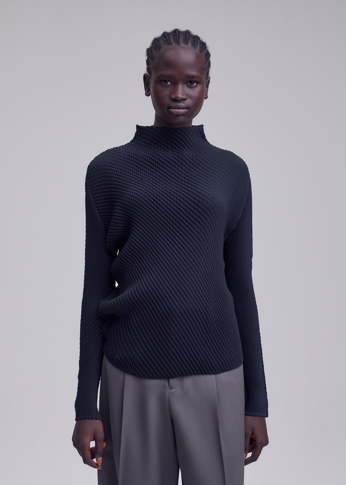 A person stands facing forward, dressed in the ISSEY MIYAKE MISTY PLEATS high-neck top with textured black fabric and loose gray trousers that create a flowy silhouette, set against a plain light gray background. view 7 of 7