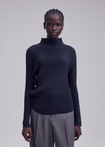 A person stands facing forward, dressed in the ISSEY MIYAKE MISTY PLEATS high-neck top with textured black fabric and loose gray trousers that create a flowy silhouette, set against a plain light gray background.