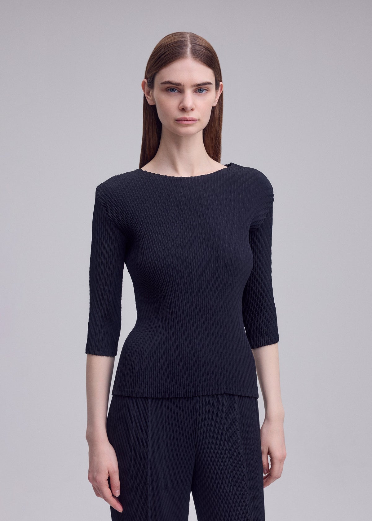 A woman with straight brown hair models the ISSEY MIYAKE RHOMBUS PLEATS, a sleek round-neck top with three-quarter sleeves and matching black pants, standing against a plain light gray background. view 5 of 5