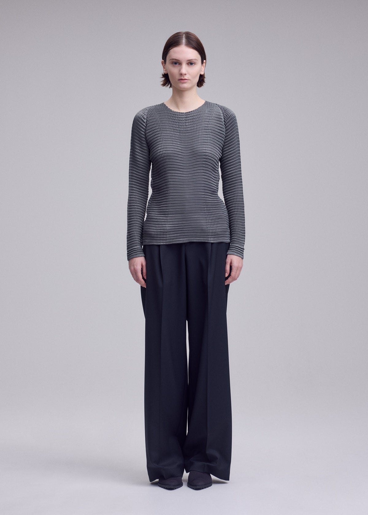Zoomed view A person faces forward in ISSEY MIYAKE’s SUEDE LIKE PLEATS: a textured gray pleated top and high-waisted, wide-leg black trousers with a suede-like finish, set against a plain light gray background.