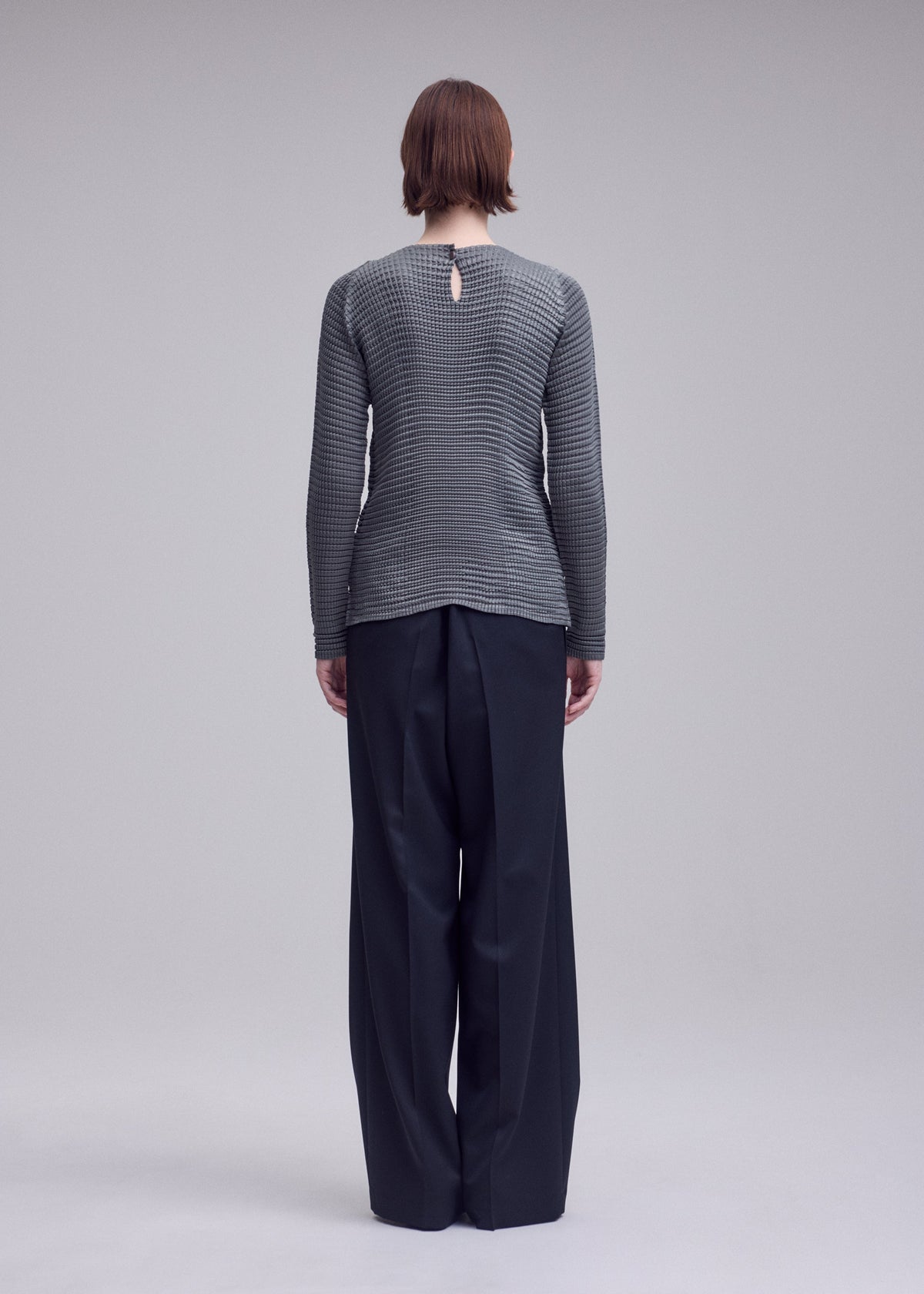 A person with short brown hair faces away, wearing the ISSEY MIYAKE SUEDE LIKE PLEATS top and loose black trousers against a plain light gray background. view 6 of 7