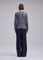 A person with short brown hair faces away, wearing the ISSEY MIYAKE SUEDE LIKE PLEATS top and loose black trousers against a plain light gray background.
