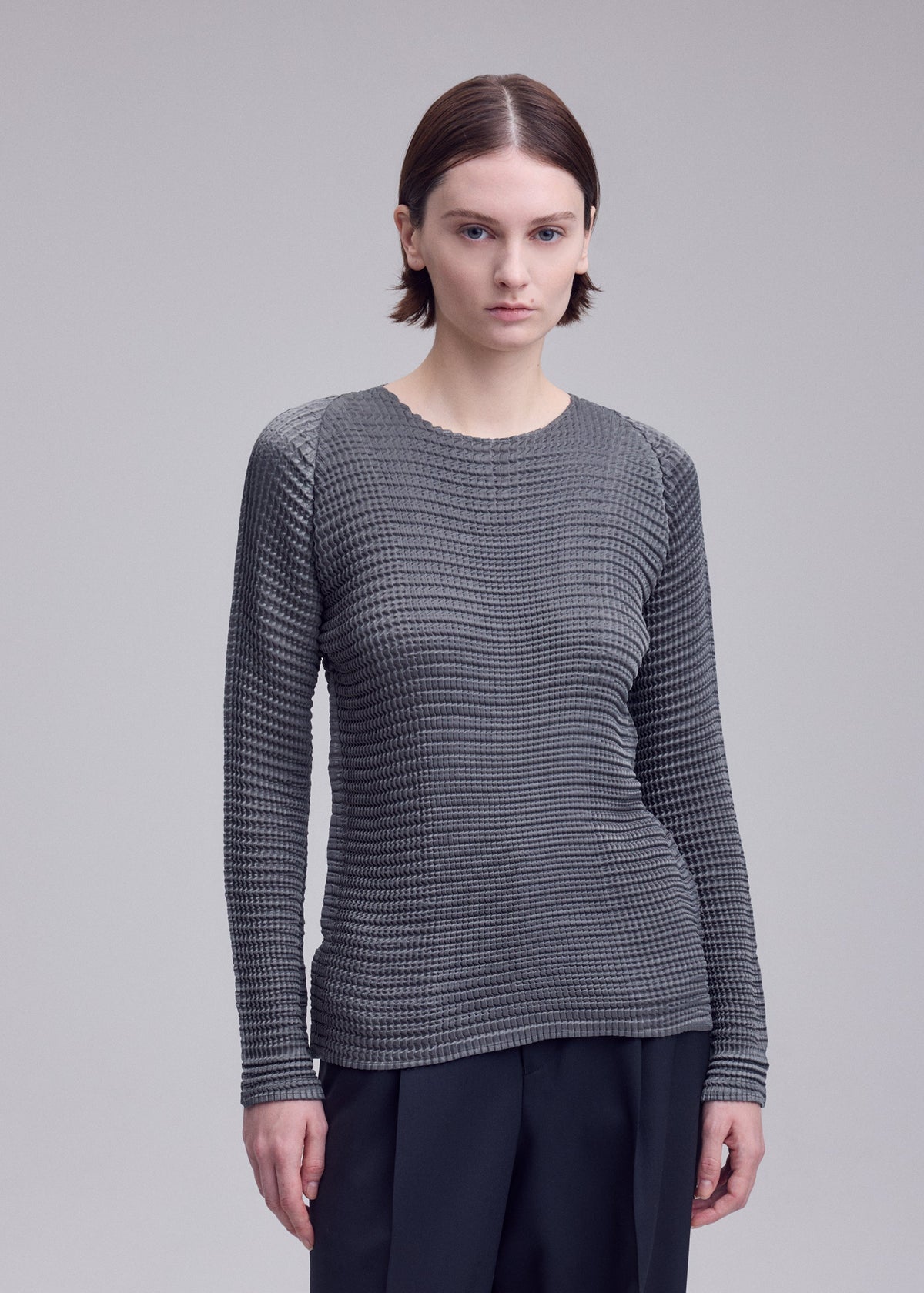 Zoomed view A person with short brown hair wears the ISSEY MIYAKE SUEDE LIKE PLEATS textured gray long-sleeve shirt and black pants, standing against a plain light gray background.
