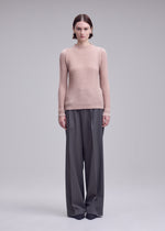 A person stands facing forward, wearing the SUEDE LIKE PLEATS top by ISSEY MIYAKE in light pink with wide-leg gray trousers, set against a plain light gray background.