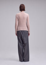 Person standing with their back to the camera, wearing the ISSEY MIYAKE SUEDE LIKE PLEATS light pink long-sleeve top and loose, high-waisted gray trousers against a plain light background.