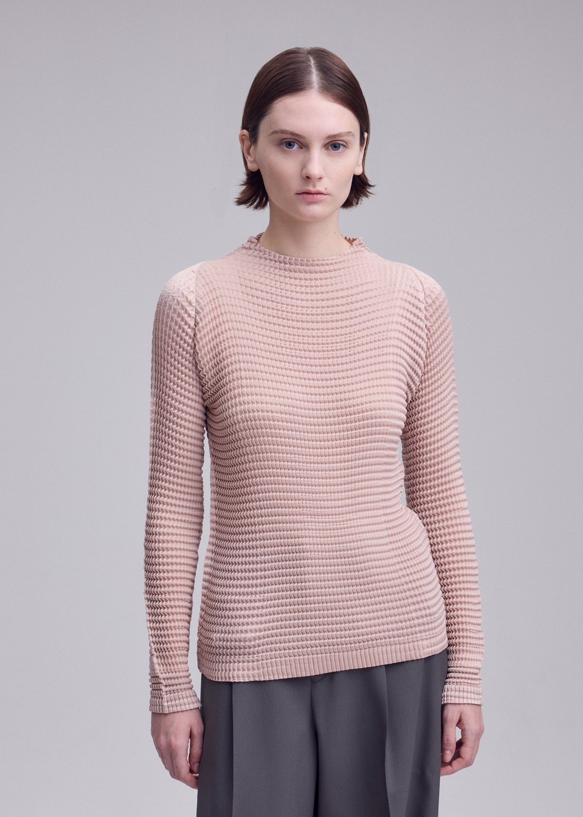 Zoomed view A person with short brown hair wears the SUEDE LIKE PLEATS by ISSEY MIYAKE— a pleated long-sleeve beige top and gray recycled polyester pants—standing against a plain light gray background.