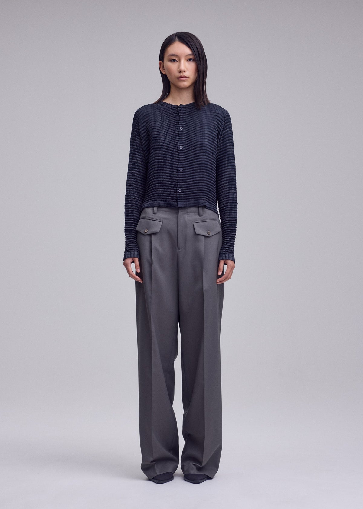A person stands facing forward in the ISSEY MIYAKE SUEDE LIKE PLEATS, a dark ribbed sweater with a suede-like sheen, paired with wide-leg gray trousers with front flap pockets against a plain light background. view 5 of 7